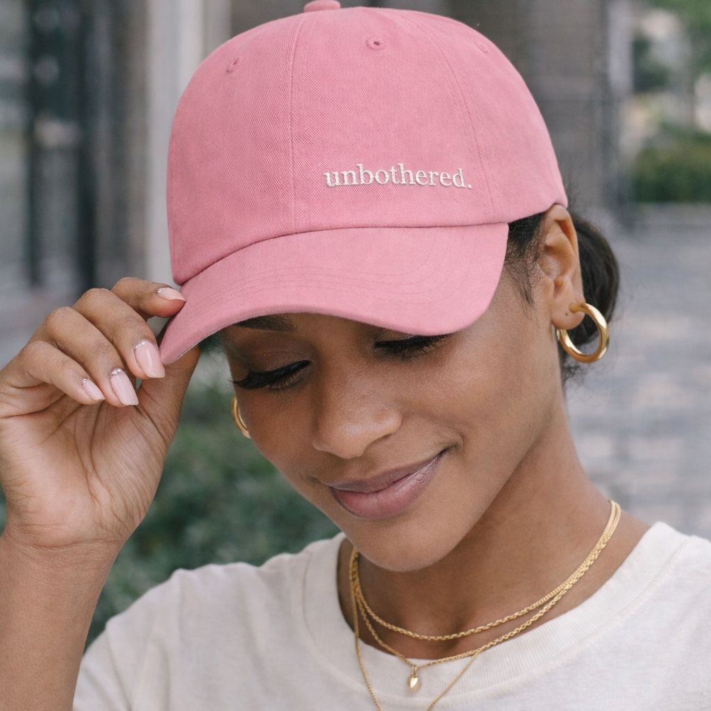 Woman wearing pink “unbothered” dad hat with minimal embroidered text, styled with neutral outfit and gold jewelry for everyday casual look