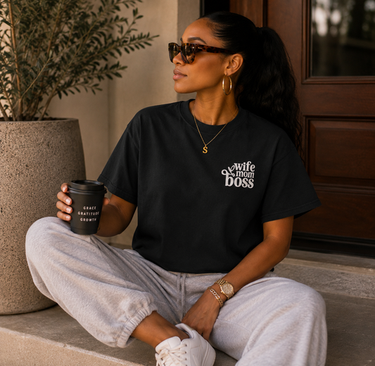 A woman sits cross-legged on outdoor steps wearing a black graphic T-shirt that reads 'wife of the boss' in white script, paired with light gray sweatpants and white sneakers. She holds a black coffee cup and wears sunglasses and gold jewelry.